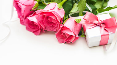Pink roses to the left of a small gift box tied with a pink ribbon.